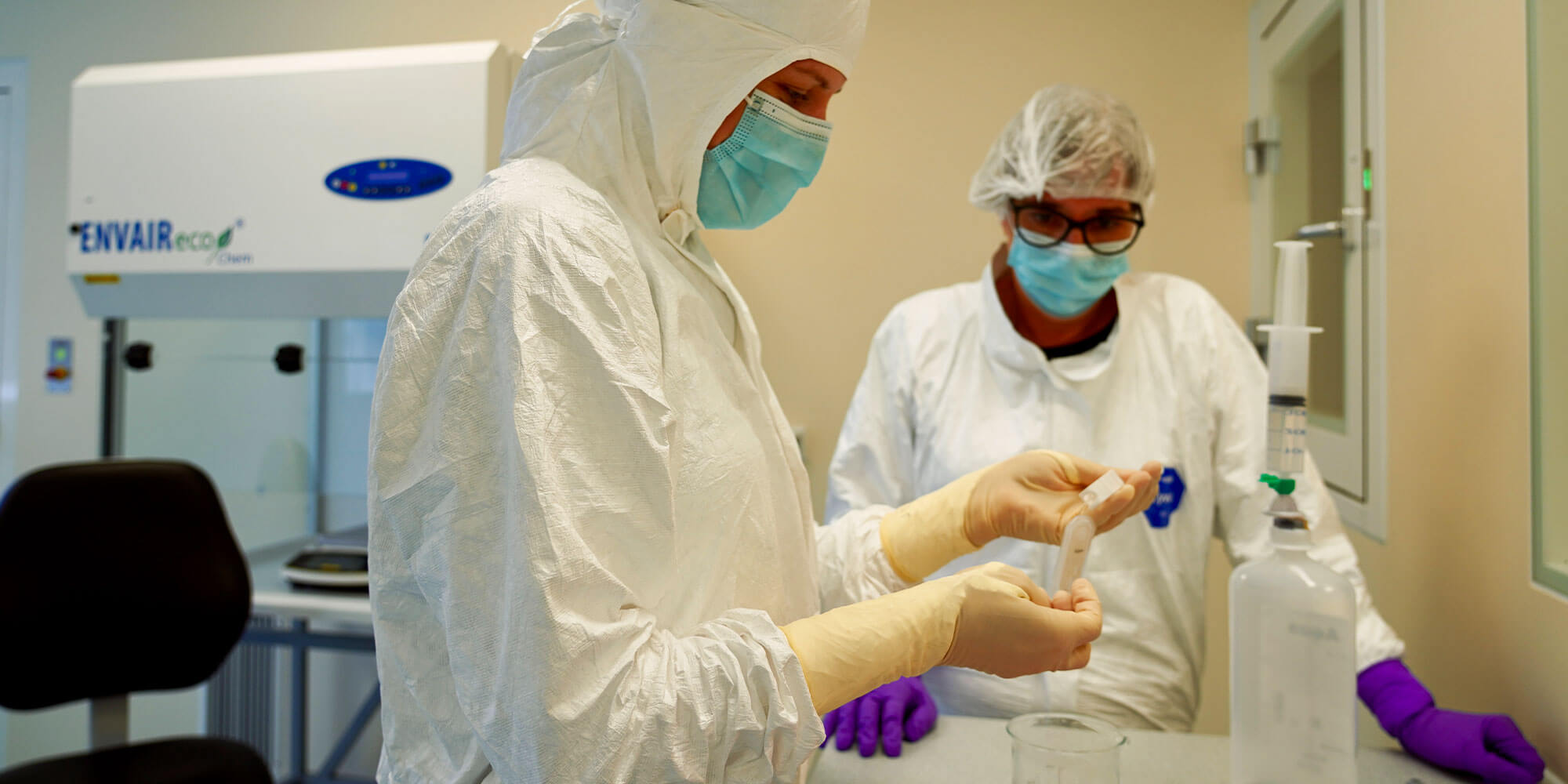 Zwei Personen in Schutzanzügen, Hauben, Masken und Handschuhen arbeiten im Lab Pharmacy Reinraumlabor mit medizinischen Proben und Infusionsmaterial.